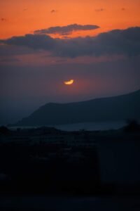 Sunset with sun partially obscured by dark clouds over silhouetted mountains and buildings.