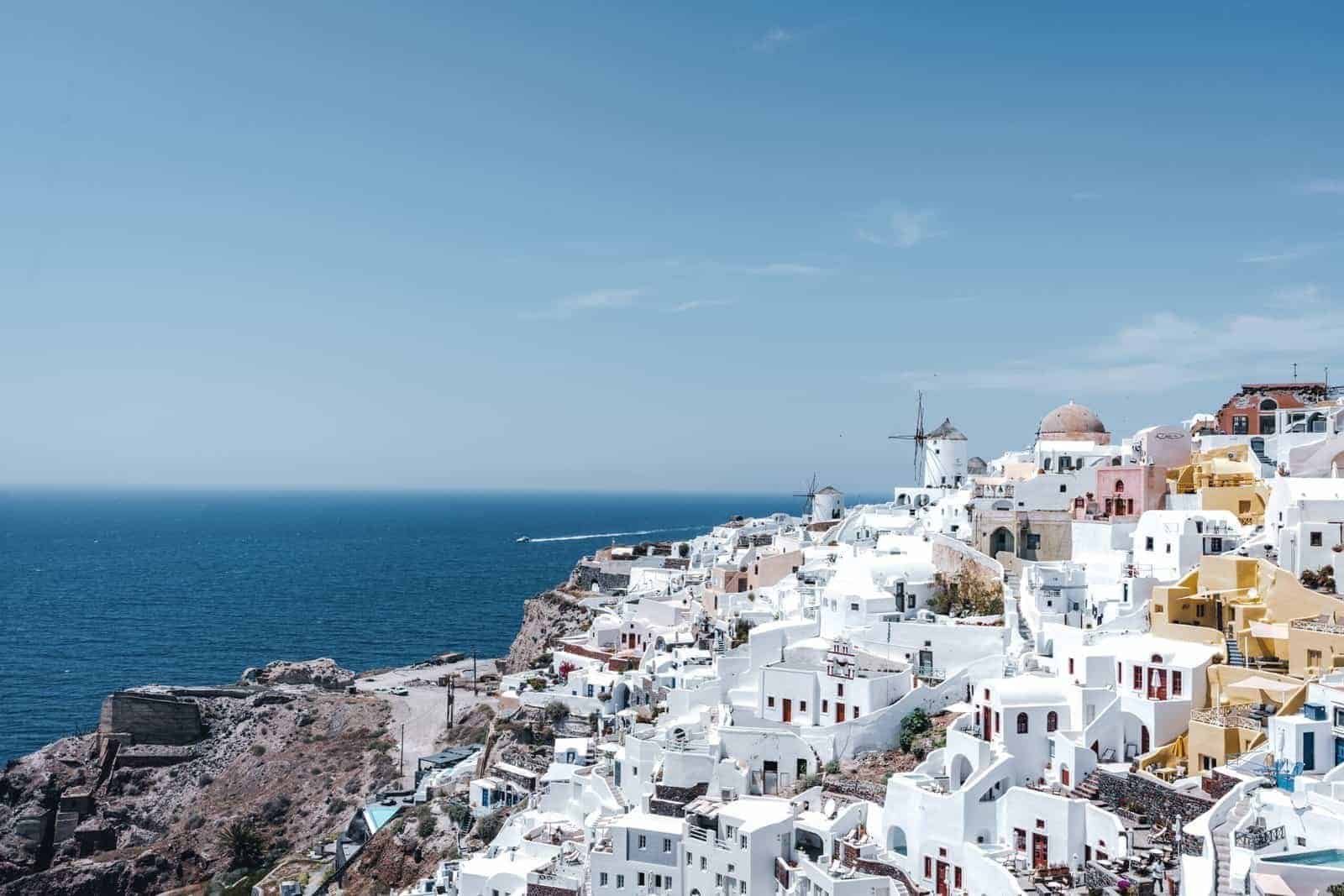 Coastal hillside town with windmill and boat trail Coastal hillside town with white and pastel buildings, windmill, and boat on deep blue sea.