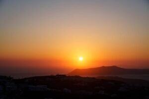 Sunset over coastal mountains with dark buildings in the foreground and orange sky gradient.