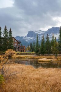 Lodge-style building by a pond with evergreen trees and snow-capped mountains under cloudy sky