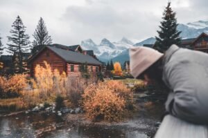 Person in a pink hat and gray jacket leaning on railing overlooking autumn landscape with cabins and mountains