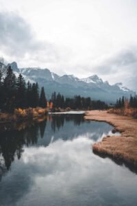 River reflecting cloudy sky with autumn trees, dry grass, and snow-capped mountains in the background.
