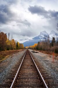 Railroad tracks extending into autumn forest with snow-capped mountains and cloudy sky.