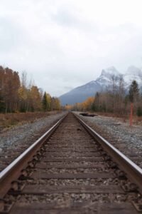 Railroad tracks extend through autumn trees toward mountains under a cloudy sky.