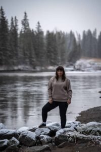 Person standing on snow-covered rocks by a river with evergreen trees in the background.