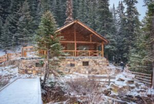 Stone and wooden cabin in snowy forest with a person walking on a fenced path nearby.