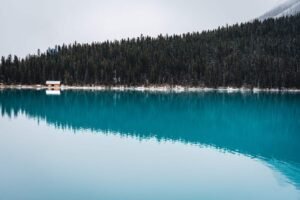 Wooden cabin by snowy forest on the edge of a turquoise lake with reflections.