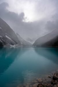 Turquoise lake with rocky shore, surrounded by misty snow-dusted mountains under cloudy sky