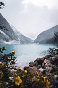 Mountain lake with turquoise water, rocky shore, yellow flowers, and snow-dusted mountains under misty sky.