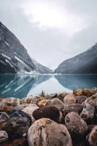 Rocky shore in foreground with a calm lake reflecting forested mountains and cloudy sky.