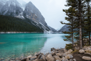 Turquoise lake with rocky shore, evergreen trees, and snow-dusted mountains under a gray sky.