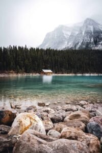 Rocky shore beside a turquoise lake with a wooden cabin, forest, and snowy mountains.