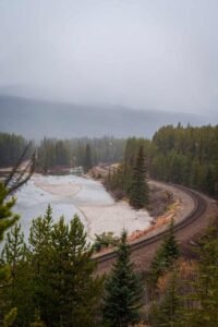 Curved railway track beside river in dense evergreen forest with snowy foggy hills.