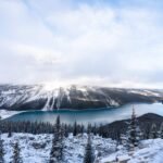 Snow-covered evergreen forest surrounds a blue lake with mist over snowy mountains.
