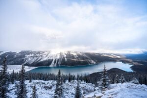 Snow-covered evergreen forest surrounds a blue lake with mist over snowy mountains.