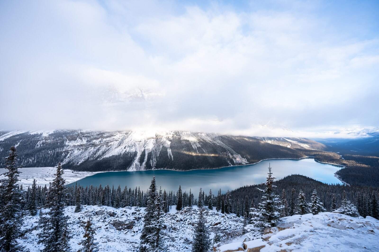 Snow-covered evergreen forest surrounds a blue lake with mist over snowy mountains.