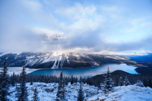 Snow-covered mountains and evergreen forest surrounding a calm blue lake under a partly cloudy sky.