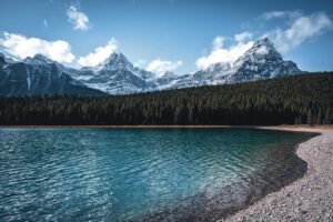 Clear blue lake with rocky shore, dense evergreen forest, and snow-capped mountains under partly cloudy sky