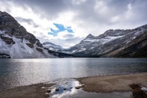 Lake surrounded by snow-dusted mountains under a partly cloudy sky with reflected rocks at shore.