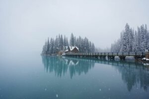 Snowy cabin and pine trees on a peninsula with a bridge over a calm lake in winter.
