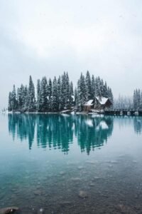 Snow-covered pine trees and houses on an island reflected in a lake with a wooden bridge.