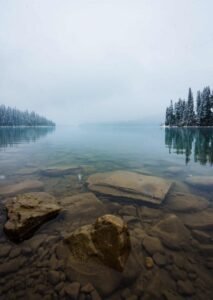 Calm lake with clear water showing submerged rocks, snow-covered pine trees, and overcast sky.
