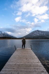 Person standing on wooden dock over lake with mountains and cloudy sky in the background