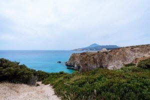 Rocky coastal cliffs with green shrubbery overlooking the blue sea and distant mountains.