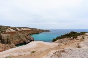 Rocky coastal cliffs surrounding a small calm bay with clear turquoise water and sparse vegetation.