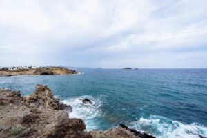 Rocky coastline with waves, blue ocean, distant white buildings, and small islands under a cloudy sky.