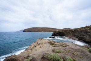 Rocky coastal cliffs with sparse vegetation by ocean waves under a cloudy sky.