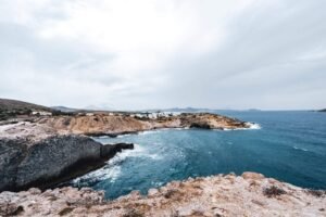 Rocky coastal cliffs with white buildings by the sea under a cloudy sky and distant hills.