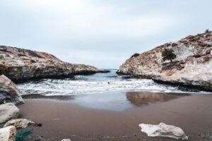 Narrow brown sand beach bordered by rocky cliffs with sparse vegetation and gentle sea waves under cloudy sky.