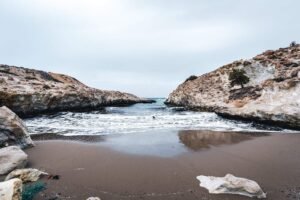 Sandy beach with waves between rocky cliffs under an overcast sky and a lone tree on right cliff.
