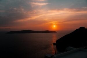 Sunset over calm water with an island in the distance and rocky silhouette on the right side.