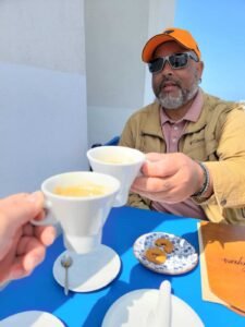 Two people toasting small white coffee cups at a blue outdoor table with cookies and a menu.