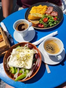 Blue table with two coffees, scrambled eggs, bacon, salad, cherry tomatoes, bread, and Greek salad with feta.