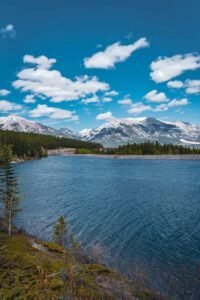 River flowing through evergreen forest with snow-capped mountains and blue sky with clouds.