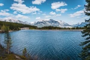 Mountain lake with forested shore under blue sky with scattered clouds.