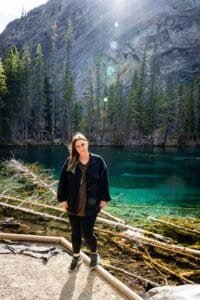 Person standing on gravel path near clear turquoise lake with fallen logs, pine trees, and rocky mountain slope.