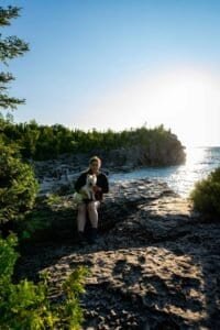 Person sitting on rocky shore holding small white dog with water and trees in background.