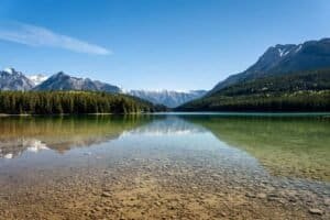 Clear shallow lake with pebble bottom, forested shores, and snow-capped mountains under blue sky.