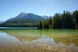 Clear lake with visible stones, surrounded by evergreen forest and mountain range under blue sky