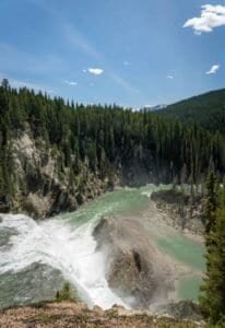 Waterfall flowing into a river surrounded by evergreen forest and distant snow-capped mountains.