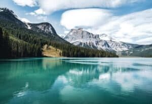 Turquoise lake with reflections of dense evergreen forests and snow-capped mountains under clouds.