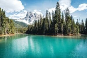 Turquoise lake surrounded by evergreen trees and snow-capped mountains under cloudy sky