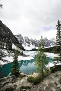 Partially frozen lake with turquoise water, surrounded by snow-covered mountains and evergreen trees.