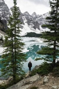 Person standing on rocky shore near a partially frozen lake with pine trees and snow mountains.