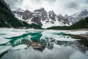Partially frozen lake reflecting snow-covered mountains and evergreen forests under cloudy sky