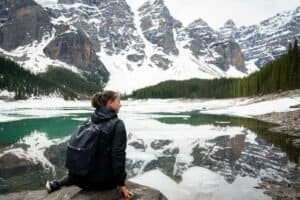 Person with backpack sitting on rock by partially frozen lake reflecting snow-covered mountains and evergreen trees.